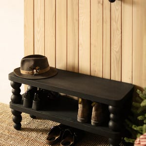May include: A black wooden shoe bench with a lower shelf and turned legs. A brown hat with a feather sits on top. Shoes and boots are stored on the shelves. Three black coat hooks are mounted on the wooden wall.