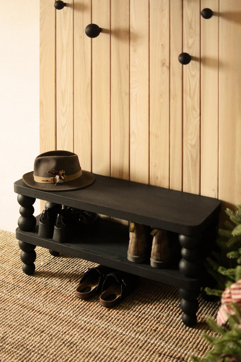 May include: A black wooden shoe bench with a lower shelf and turned legs. A brown hat with a feather sits on top. Shoes and boots are stored on the shelves. Three black coat hooks are mounted on the wooden wall.