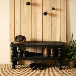 May include: A black shoe bench with a lower shelf and turned legs. A brown hat sits on top, with shoes and boots on the shelves. A wooden wall panel with black round hooks is behind the bench. A small potted evergreen tree is to the right.