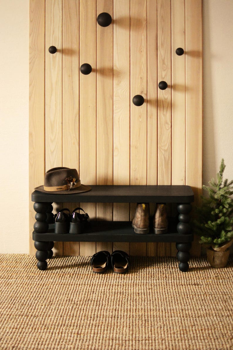 May include: A black shoe bench with a lower shelf and turned legs. A brown hat sits on top, with shoes and boots on the shelves. A wooden wall panel with black round hooks is behind the bench. A small potted evergreen tree is to the right.