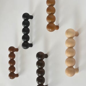 May include: A collection of wooden cabinet pulls in various shades of brown and black. Each pull features a series of connected spheres, creating a unique and modern design. The pulls are mounted on a white surface, showcasing their different wood tones.
