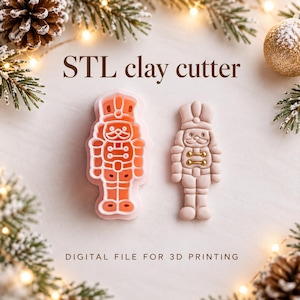Könnte beinhalten: Ein hellrosa Ausstechförmchen in Nussknacker-Form und eine entsprechende geformte Tonfigur. Der Text lautet STL clay cutter und DIGITAL FILE FOR 3D PRINTING. Die Nussknacker-Figur hat kleine goldfarbene Akzente auf der Brust.