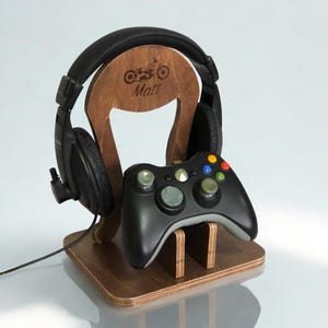 May include: Wooden headphone stand with a motorcycle graphic and the name "Matt" engraved. A black gaming controller rests on the stand, with black headphones hanging from the top. The stand is made of light brown wood.