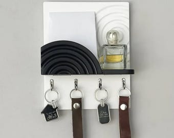 May include: White wall mounted key holder with a black and white design. It has a small shelf for mail or other items and four hooks for keys. There is a small bottle of perfume on the shelf. The hooks have keychains with house and dog tags.