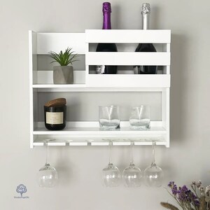May include: White wall mounted wine rack with two shelves, a glass holder, and a small plant on the top shelf. The rack is holding two bottles of wine and two glasses.
