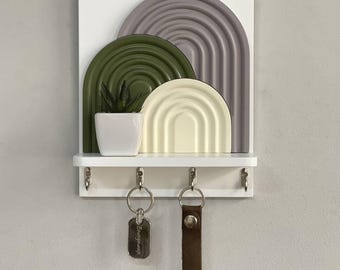 May include: A white wall-mounted key holder with a shelf and three hooks. It features a decorative design of three arched shapes in olive green, cream, and gray. A small potted plant sits on the shelf, and two sets of keys hang from the hooks.