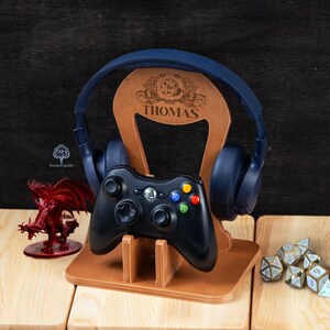 May include: A wooden gaming headset and controller stand engraved with the name "THOMAS". The stand supports a black Xbox controller and blue headphones. A red dragon figurine and dice are also present.