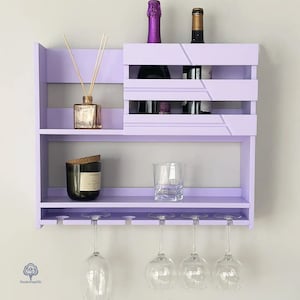 May include: A lavender wall mounted wine rack with two shelves, a glass holder, and a decorative bottle holder. The top shelf has a decorative bottle and a diffuser. The bottom shelf has a candle and a glass.
