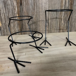 May include: Three black metal plant stands with circular holders and tripod legs. The stands vary in height and size, with a minimalist design. The background is a wooden surface.