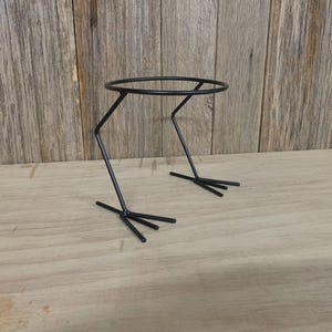 Può includere: Un portapiante in metallo nero con una parte superiore circolare e gambe progettate per assomigliare a zampe di rana. Il supporto è realizzato con sottili aste di metallo ed è adatto per contenere piccole piante in vaso o oggetti decorativi. Il supporto è alto circa 15 cm.