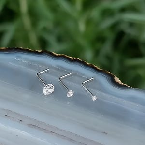 May include: Three silver nose rings with clear cubic zirconia stones. The stones are different sizes, with the largest stone in the centre.