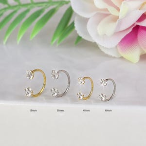 May include: Four horseshoe-shaped body jewellery pieces featuring clear round-cut stones at each end. Two gold-tone pieces are labelled 8mm and 6mm, and two silver-tone pieces are labelled 8mm and 6mm. The jewellery is arranged in a row on a white surface.