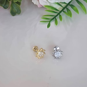 May include: Two small, silver and gold, sun-shaped earrings with a circle of clear rhinestones in the centre. The earrings are on a white surface.
