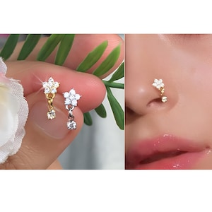 May include: A pair of nose rings featuring a flower design with a dangling crystal. The rings are shown on a hand and on a person's nose.