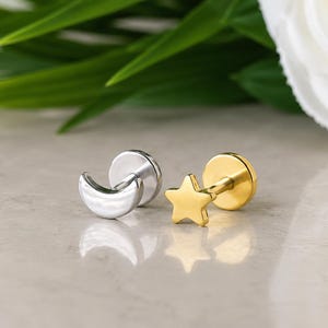 May include: A silver-toned crescent moon stud earring and a gold-toned star stud earring. Both earrings feature a flat circular backing and a post design, showcasing a polished metallic finish on the moon and star shapes respectively.