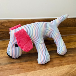 May include: A striped plush toy dog with pink ears and a black eye. The dog is made of fabric and has a white background with colourful stripes.