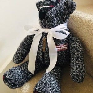 May include: A black and white knitted teddy bear with a white ribbon around its neck. The bear has a red and white plaid patch on its foot and a tag that reads "Tommy Edmonds 1988-2009".