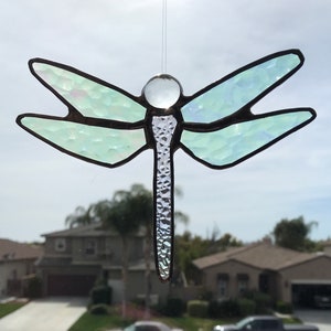 May include: A stained glass dragonfly suncatcher with iridescent wings and a textured body. The dragonfly is suspended by a thin wire, with a clear glass bead at the top. The wings are a light green color.