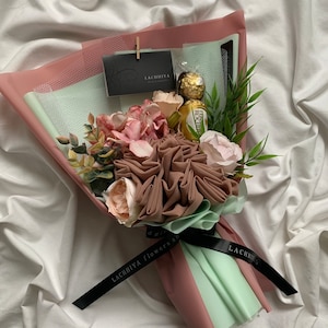 May include: A bouquet wrapped in pink and mint green paper, with fabric flowers in pink and brown, greenery, and a gold-wrapped chocolate. A black ribbon with the text "LACHHIYA flowers and..." is tied around the bouquet.