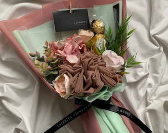 May include: A bouquet wrapped in pink and mint green paper, featuring fabric flowers in shades of pink and brown, with greenery and a gold-wrapped chocolate. A black ribbon with the text "LACHHIYA flowers and..." is tied around the bouquet.