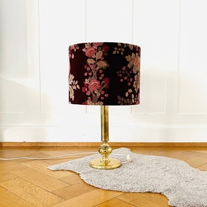Könnte beinhalten: Eine goldene Tischlampe mit einem floralen Lampenschirm. Der Lampenschirm ist in einem tiefen Burgunderrot mit rosa und cremefarbenen floralen Akzenten gehalten. Die Lampe steht auf einem weißen, flauschigen Teppich auf einem Holzboden.