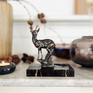 May include: A bronze-colored deer figurine standing on a black marble base with gold veining. The deer is posed with its head up and antlers visible. The figurine is a decorative item, likely for a home or office.