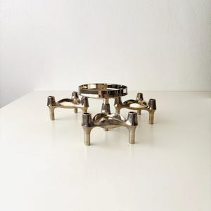Peut inclure: Un bougeoir en métal argenté composé d'une plateforme circulaire centrale surélevée sur un piédestal, entourée de quatre bras porte-bougies individuels, chacun soutenu par trois pieds fins, formant une structure interconnectée à plusieurs niveaux.