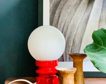 May include: A red and white table lamp with a white globe shade. The lamp is on a wooden table with two wooden candle holders.