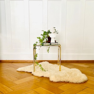 Peut inclure: Une table d'appoint en métal doré avec un plateau en verre et un tapis en fausse fourrure blanche. Une plante verte en pot et un buste blanc sont sur la table.