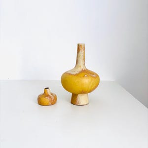 Könnte beinhalten: Zwei gelbe Keramikvasen mit weißem Überzug. Die größere Vase hat einen breiten, abgerundeten Boden und einen schmalen Hals. Die kleinere Vase hat eine ähnliche Form, ist aber viel kleiner.