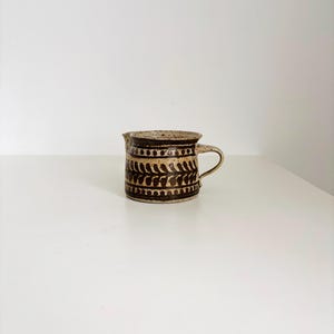 Op de afbeelding: Een kleine, handgemaakte keramische kan met een donkerbruin en beige kleurenschema. De kan heeft een decoratief patroon van bladvormige vormen en een klein handvat. De rand en het handvat zijn beige, terwijl de body donkerbruin is.