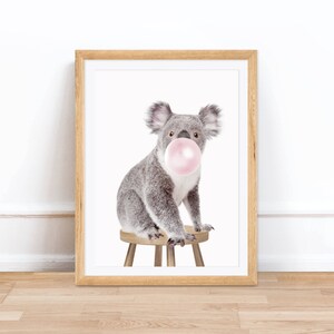 Op de afbeelding: Een ingelijste print met een grijze koala die op een kleine houten kruk zit. De koala heeft een grote roze kauwgomballenbel in zijn mond. De print zit in een lichtbruine houten lijst, ideaal voor een kinderkamer.