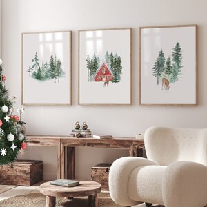May include: Three watercolour paintings in wooden frames depicting a winter forest scene. The centre painting shows a red cabin with a snowy roof and trees in the background. The left and right paintings show a deer in the forest.