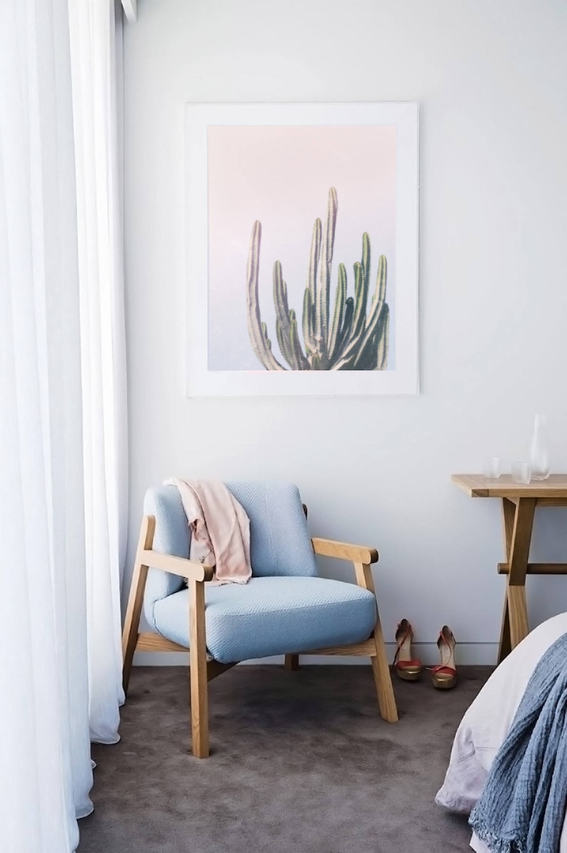 May include: A light blue armchair with wooden legs sits in a room with white walls and a framed print of a cactus against a pink background.
