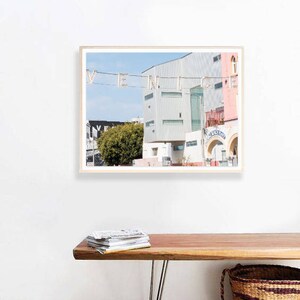 May include: A framed photograph of a building with the word "VENICE" spelled out in lights. The building is painted in shades of pink and white, and there is a green tree in the foreground. The photograph is hanging on a white wall above a wooden bench with a stack of magazines on it.