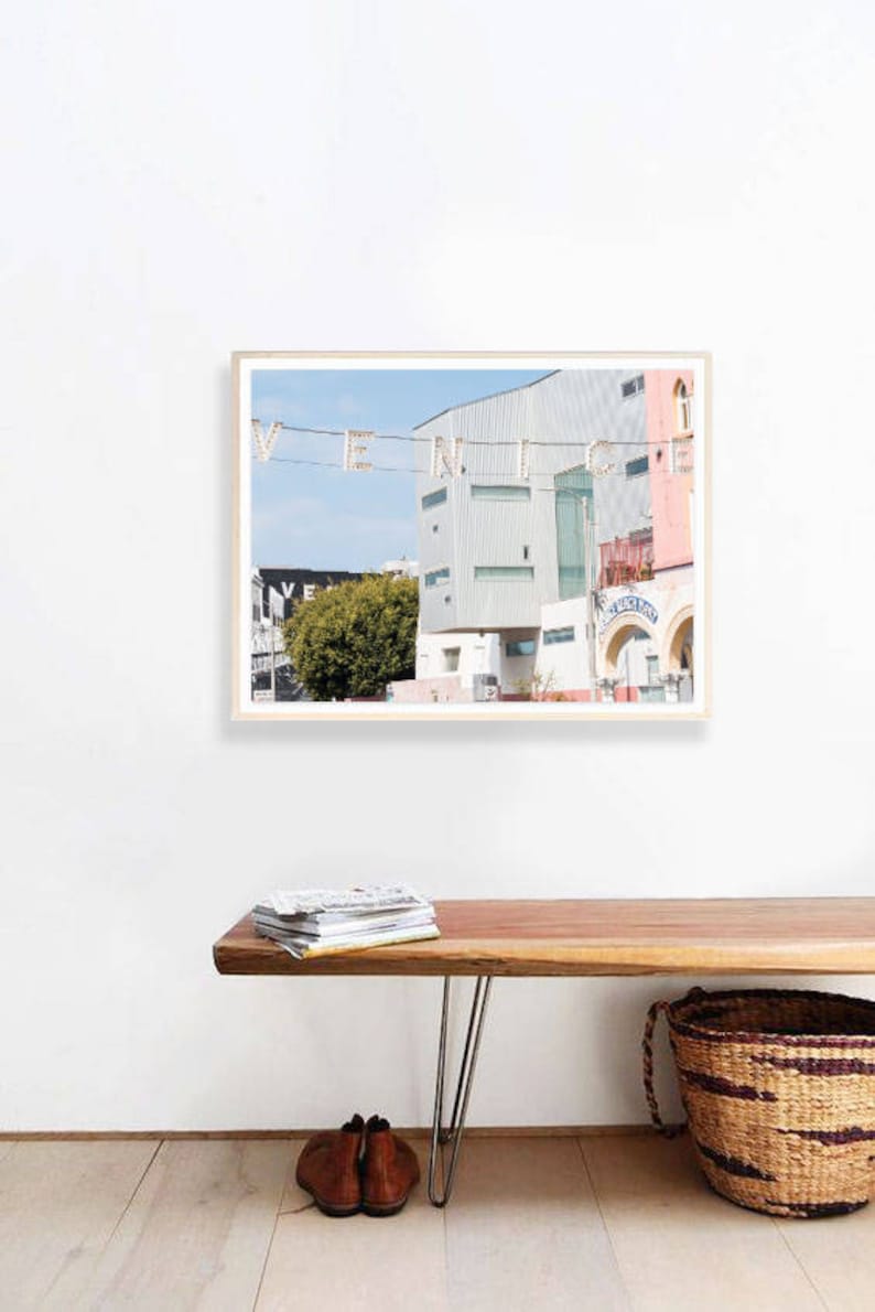 May include: A framed photograph of a building with the word "VENICE" spelled out in lights. The building is painted in shades of pink and white, and there is a green tree in the foreground. The photograph is hanging on a white wall above a wooden bench with a stack of magazines on it.