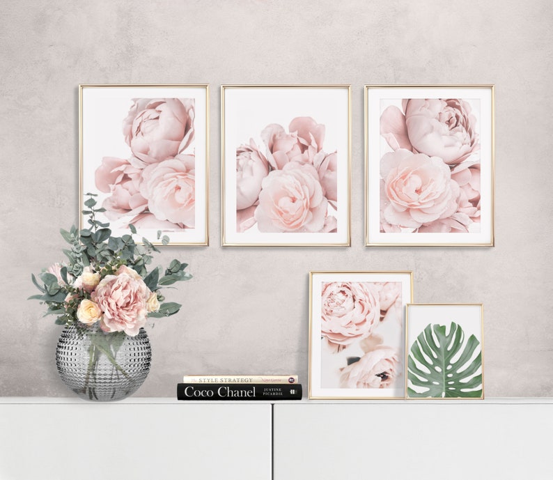 Op de afbeelding: Vier ingelijste prints van roze pioenrozen met gouden lijsten, een boek getiteld "Coco Chanel" en een vaas met roze bloemen op een wit oppervlak.