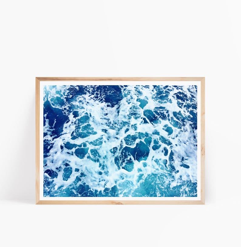 May include: A close-up photograph of blue ocean water with white foam and bubbles. The image is framed in a light wood frame.