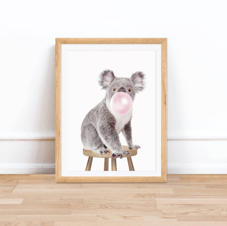 Op de afbeelding: Een ingelijste print met een grijze koala die op een kleine houten kruk zit. De koala heeft een grote roze kauwgomballenbel in zijn mond. De print zit in een lichtbruine houten lijst, ideaal voor een kinderkamer.