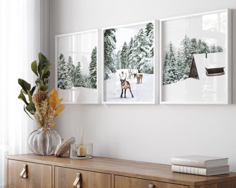 May include: Three framed winter landscape prints with white frames. The center print shows a snowy forest with reindeer. The left print shows a snowy forest. The right print shows a snowy forest with a small cabin.