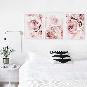 May include: Three framed prints of pink roses, arranged in a row on a white wall above a white bed with a black and white patterned pillow.