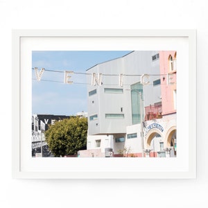 May include: A photograph of a street scene in Venice, California. The word "VENICE" is spelled out in white lights strung across the street. There are buildings in the background, including one with a sign that says "Venice Beach Market".