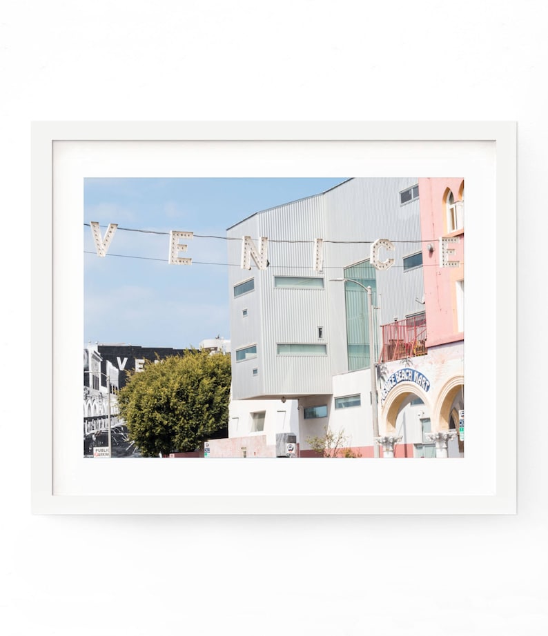 May include: A photograph of a street scene in Venice, California. The word "VENICE" is spelled out in white lights strung across the street. There are buildings in the background, including one with a sign that says "Venice Beach Market".