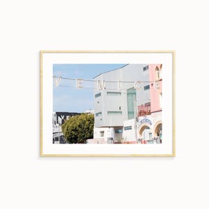May include: A photograph of a street scene in Venice, California. The word "VENICE" is spelled out in large, white letters strung across the street. The letters are illuminated with small, white lights. The photo is taken from a low angle, looking up at the buildings. The buildings are a mix of modern and traditional styles. The sky is blue and there are clouds in the distance.