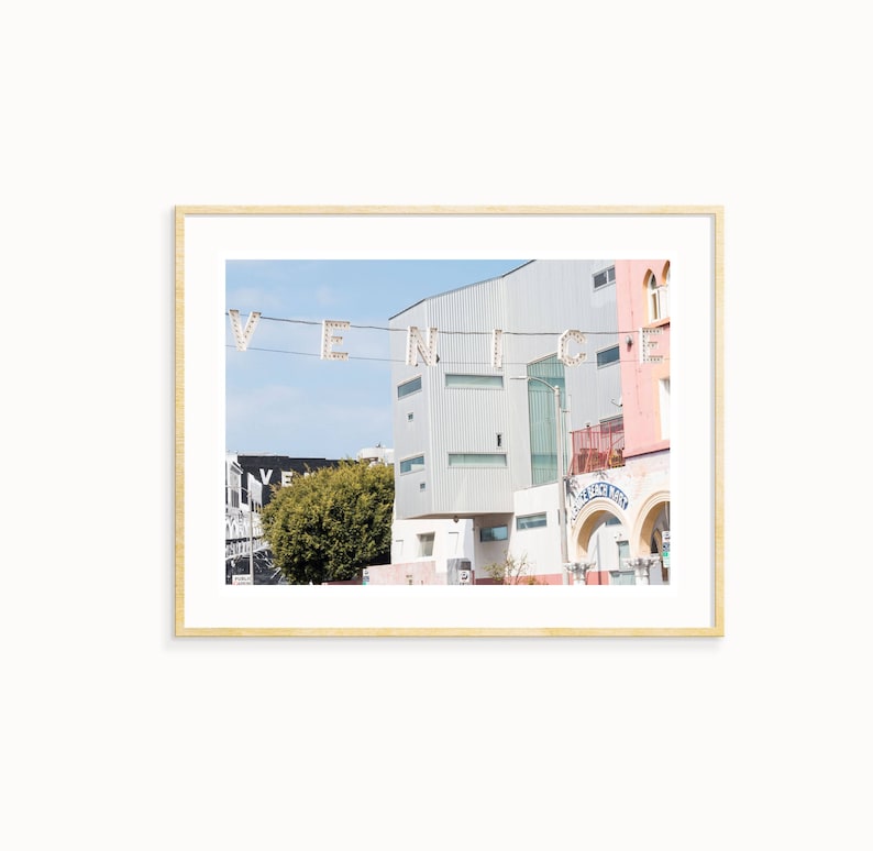 May include: A photograph of a street scene in Venice, California. The word "VENICE" is spelled out in large, white letters strung across the street. The letters are illuminated with small, white lights. The photo is taken from a low angle, looking up at the buildings. The buildings are a mix of modern and traditional styles. The sky is blue and there are clouds in the distance.