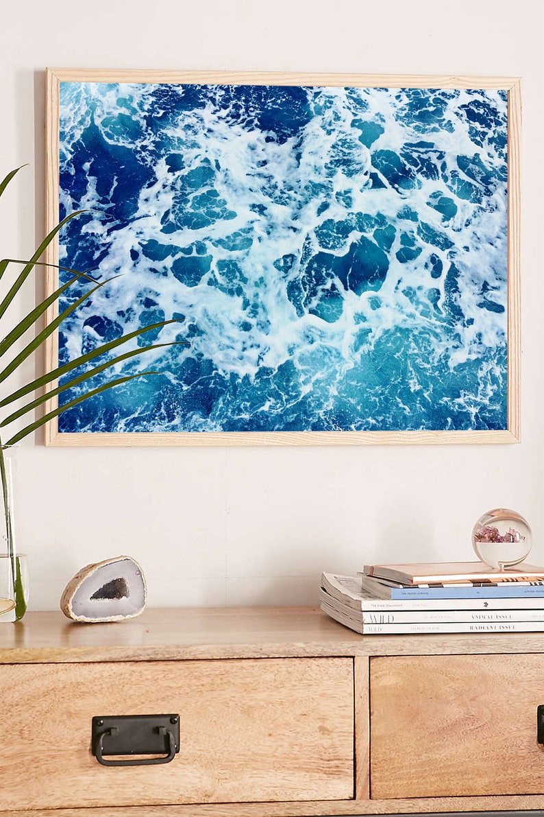 May include: A framed photograph of a blue ocean with white foam waves. The photo is in a light wood frame and is hanging on a white wall.