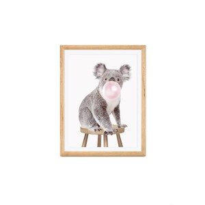 Op de afbeelding: Een ingelijste print met een grijze koala die een roze kauwgomballenblaas blaast. De koala zit op een kleine houten kruk. De lijst is gemaakt van lichtbruin hout, tegen een witte achtergrond.