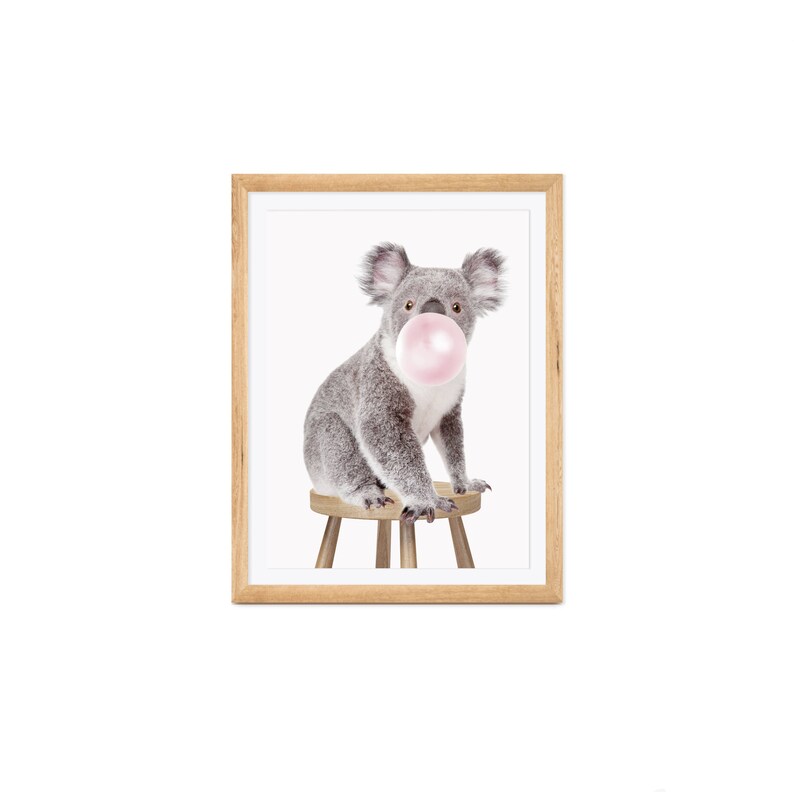 Op de afbeelding: Een ingelijste print met een grijze koala die een roze kauwgomballenblaas blaast. De koala zit op een kleine houten kruk. De lijst is gemaakt van lichtbruin hout, tegen een witte achtergrond.