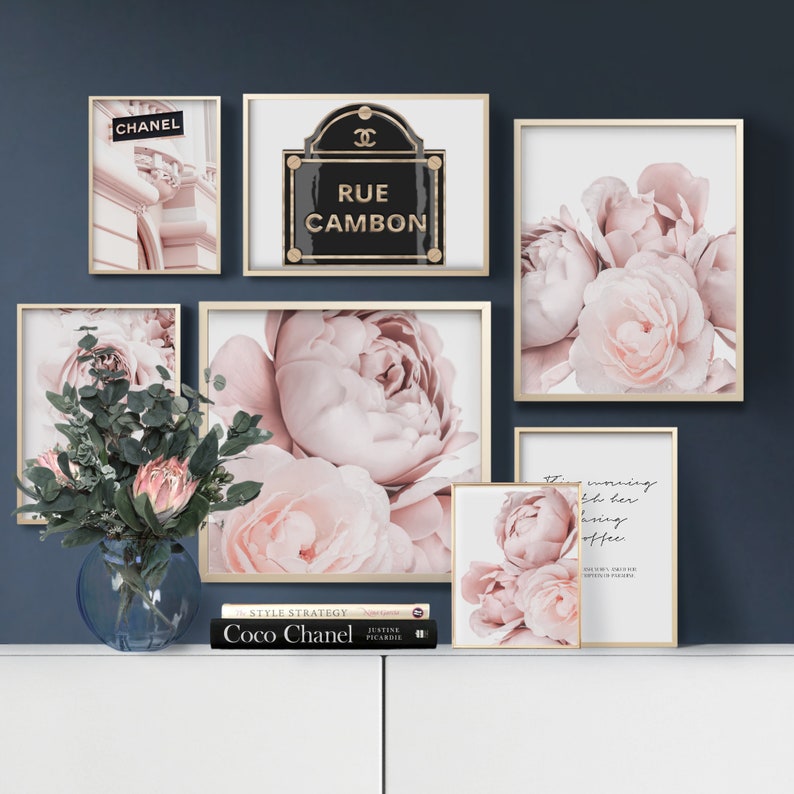 Op de afbeelding: Een set van zes ingelijste prints met roze pioenen en een zwart en gouden bord met de tekst "Rue Cambon". De prints zijn gerangschikt op een muur met een blauwe achtergrond. Een boek met de titel "Coco Chanel" staat op een witte plank onder de prints.