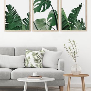 May include: Three framed prints of tropical leaves in shades of green against a white background. The prints are arranged in a row on a white wall.